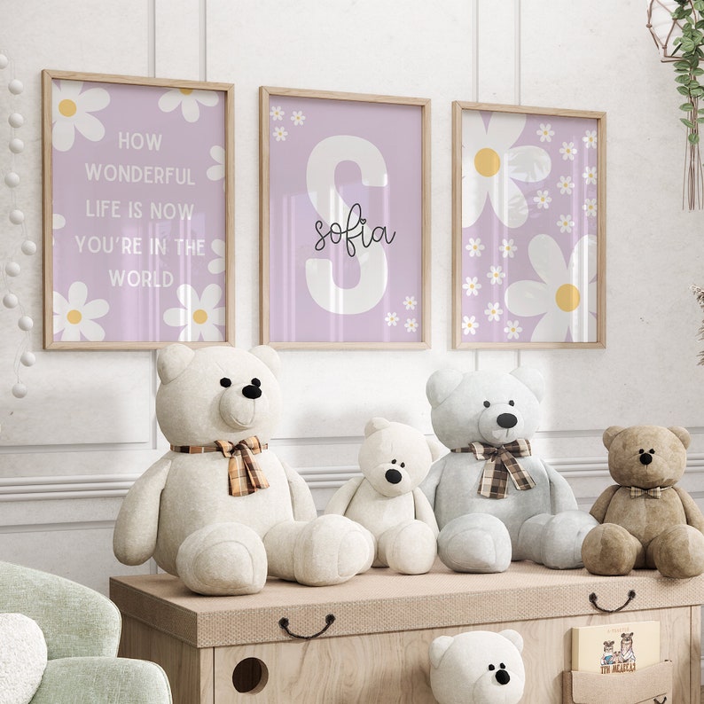 May include: Three framed prints with a purple background and white daisy flowers. The center print has a large white letter "S" with the name "Sofia" written in black cursive. The prints are hanging on a white wall above a wooden shelf with four teddy bears.