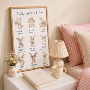 May include: A framed print with the text "GOD SAYS I AM" and illustrations of bunnies, along with inspirational words. A small lamp, a mug with hearts, a vase with flowers, and a book are on a white surface. Pink pillows and bedding are in the background.