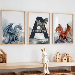 May include: Three framed art prints featuring watercolor dragons. One dragon is blue, one is red, and the third has a green dragon with the name "ANDREW" in white text. The prints have a light wood frame.
