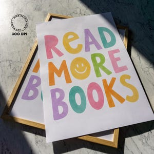 May include: Two framed art prints with the words "READ MORE BOOKS" in colorful, playful lettering. The text includes a yellow smiley face and is set against a white background. The frames are a light wood color.