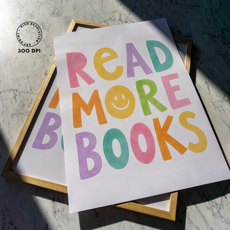 May include: Two framed art prints with the words "READ MORE BOOKS" in colorful, playful lettering. The text includes a yellow smiley face and is set against a white background. The frames are a light wood color.