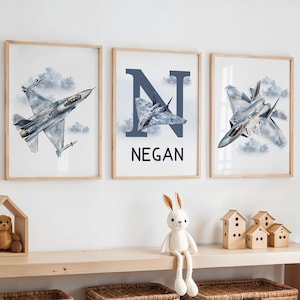 May include: Three framed prints with watercolour illustrations of fighter jets against a cloudy sky. The central print shows the name "NEGAN" below a stylized letter "N" and a jet illustration. The frames are light wood.
