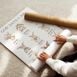 May include: A rolled-up poster with the text "GOD SAYS I AM" and illustrations of bunnies with floral accents. The poster includes words like "LOVED", "BLESSED", "CHOSEN", and "UNIQUE". A brown cardboard tube rests on top.