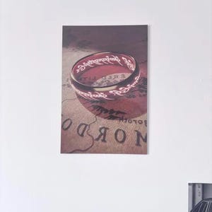 May include: A rectangular wall print featuring a gold-toned ring with glowing, etched Elvish script. The ring is positioned over a vintage-style map with visible text and cartographic lines, creating a detailed composition of fantasy-themed imagery.