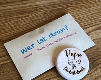Könnte beinhalten: Eine Holz-Münze mit dem Text "Wer ist dran? Mama / Papa Entscheidungsmunze" auf der einen Seite und "Papa Du bist dran!" mit einem Cartoon-Baby auf der anderen Seite.