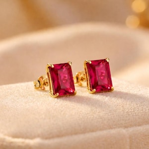 May include: A pair of gold stud earrings with rectangular, deep red gemstones. The earrings are set in a gold frame and rest on a soft, beige surface. The background is a blurred, warm tone.