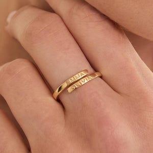以下が含まれることがあります： 2つの重なり合うバンドが特徴のゴールドトーンのバイパスリング。各バンドには名前が刻印されており、上のバンドには「EMMA」、下のバンドには「OLIVIA」という名前が、明瞭な大文字のサンセリフ体で記されています。