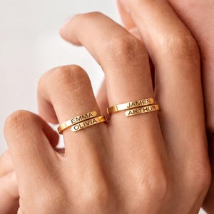 Könnte beinhalten: Zwei goldfarbene Metallringe an den Fingern. Ein Ring zeigt die eingravierten Namen EMMA und OLIVIA auf getrennten, übereinanderliegenden Bändern. Der andere Ring zeigt die eingravierten Namen JAMES und ARTHUR auf getrennten Bändern.