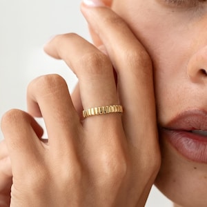 May include: A gold ring featuring a repeating vertical ribbed or fluted texture around the entire band. The ring has a polished, metallic finish and a consistent width, worn on a finger with the hand positioned near the side of the face.