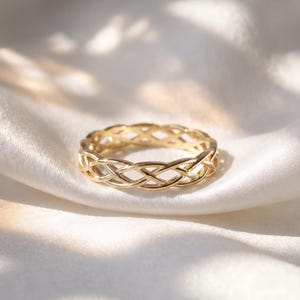 Op de afbeelding: Een gouden ring met een ingewikkeld gevlochten of geweven metaalontwerp dat de gehele band omcirkelt. De ring heeft een gepolijste afwerking en een doorlopend patroon van in elkaar grijpende lussen, wat zorgt voor een gedetailleerde textuur.