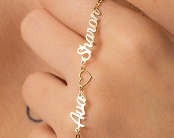 May include: A delicate gold-colored bracelet featuring the names "Ava" and "Sharon" in a vertical arrangement, separated by a small heart-shaped charm. The bracelet is held by a person, showcasing its fine chain and personalized design.