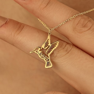 May include: A gold-colored hummingbird pendant necklace. The pendant is a detailed cutout of a hummingbird with its wings spread. The necklace is on a delicate chain.