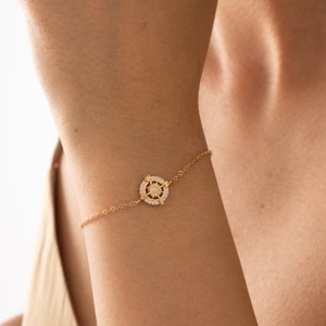 May include: A gold bracelet with a compass charm featuring a circle of small clear stones around a larger clear stone in the center.