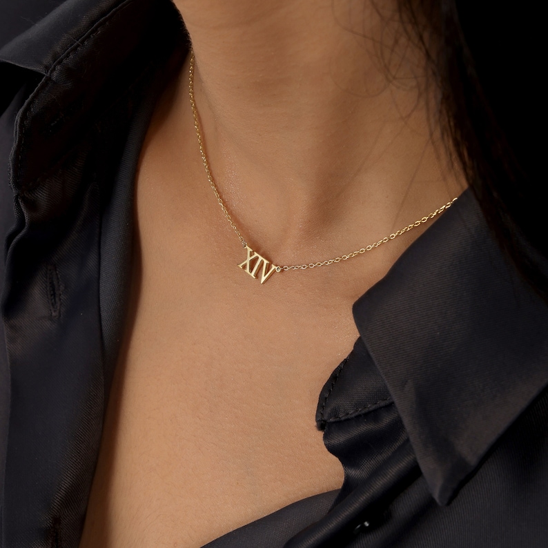 May include: A gold necklace with a delicate chain and a small pendant featuring the Roman numeral XIV.