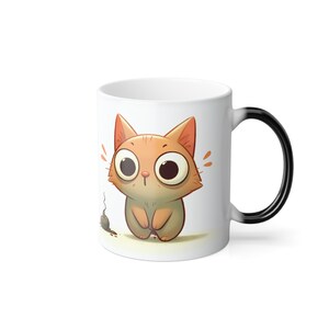 Puede incluir: Taza de cerámica blanca con un gato naranja de dibujos animados con ojos grandes y una expresión de sorpresa. También hay un pequeño ratón. El asa de la taza es negra.