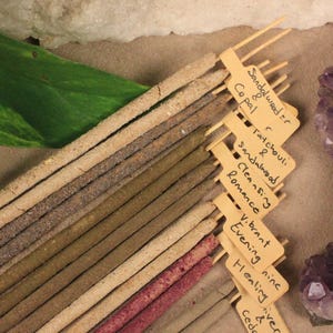 May include: A selection of incense sticks in different shades, with small labels displaying scents such as sandalwood and copal. A large purple amethyst crystal and a green leaf are also present.