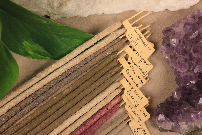 May include: A selection of incense sticks in different shades, with small labels displaying scents such as sandalwood and copal. A large purple amethyst crystal and a green leaf are also present.