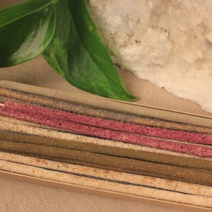 May include: A selection of incense sticks in a box, with shades of pink, brown, and beige. The sticks have small labels with text, including "Patcho & Sandal" and "Lavender Feminine Healing Jar". A green leaf and a white stone are in the background.