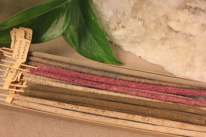 May include: A selection of incense sticks in a box, with shades of pink, brown, and beige. The sticks have small labels with text, including "Patcho & Sandal" and "Lavender Feminine Healing Jar". A green leaf and a white stone are in the background.