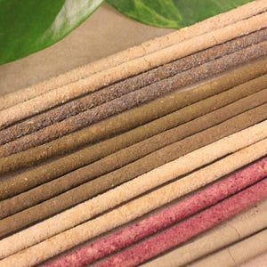 May include: A close-up shot of several incense sticks in shades of brown, beige, and pink. The sticks are arranged diagonally, with a green leaf in the top left corner. The incense sticks have a textured, granular appearance.