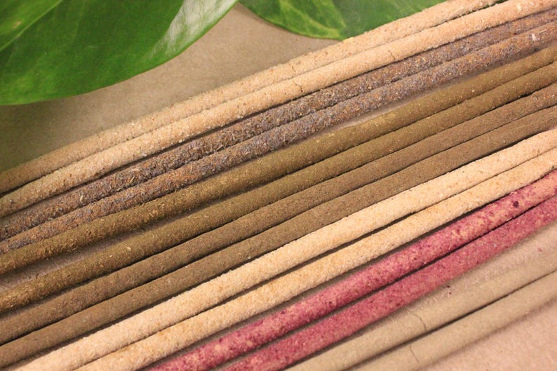 May include: A close-up shot of several incense sticks in shades of brown, beige, and pink. The sticks are arranged diagonally, with a green leaf in the top left corner. The incense sticks have a textured, granular appearance.