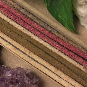 May include: Incense sticks in a wooden tray, with shades of brown, pink, and beige. The image includes purple amethyst crystals, a green leaf, and a white crystal. The incense sticks are arranged in parallel lines.