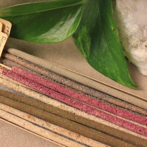 May include: A selection of incense sticks in a box, featuring shades of pink, brown, and beige. The labels read "Patchouli & Sandalwood" and "Lavender Feminine Healing Bar". Green leaves and a white crystal are also visible in the image.