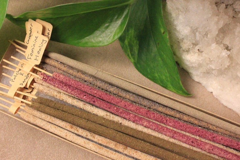May include: A selection of incense sticks in a box, featuring shades of pink, brown, and beige. The labels read "Patchouli & Sandalwood" and "Lavender Feminine Healing Bar". Green leaves and a white crystal are also visible in the image.