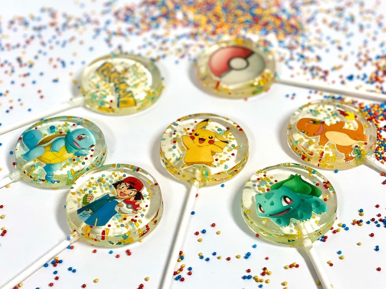 May include: Six round lollipops with different Pokemon characters on them. The lollipops are clear and have colorful sprinkles on them. The lollipops are on a white surface with colorful sprinkles scattered around them.