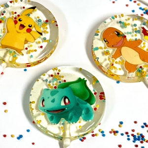 May include: Three round lollipops with colorful sprinkles. Each lollipop features a different Pokemon character: Pikachu, Charmander, and Bulbasaur.