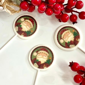 Könnte beinhalten: Drei weiße Weihnachts-Lollipop-Dekorationen mit einem Weihnachtsmann-Design. Jeder Lollipop zeigt ein rundes Bild des Weihnachtsmanns in rotem Anzug und weißem Bart, umgeben von einem grünen und roten Rand. Rote Beeren und Grünzeug sind im Hintergrund.