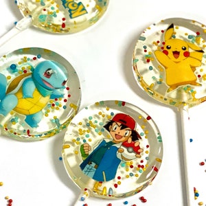 May include: Four round lollipops with colorful sprinkles and cartoon images of Pokemon characters. The lollipops are on a white background with red, yellow, and blue sprinkles.