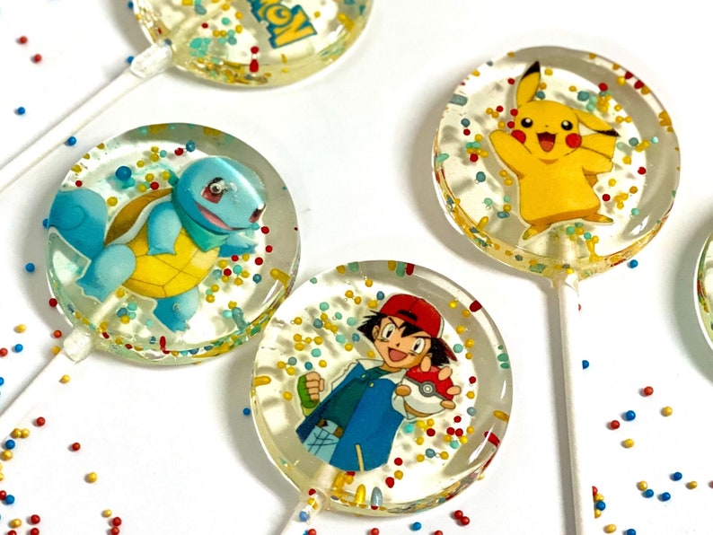 May include: Four round lollipops with colorful sprinkles and cartoon images of Pokemon characters. The lollipops are on a white background with red, yellow, and blue sprinkles.