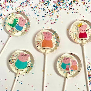 Könnte beinhalten: Fünf runde Lutscher mit bunten Streuseln auf weißem Hintergrund. Jeder Lutscher zeigt einen anderen Charakter aus der Zeichentrickserie Peppa Pig. Die Charaktere sind Peppa, George, Papa Pig, Mama Pig und Peppas Dinosaurier.