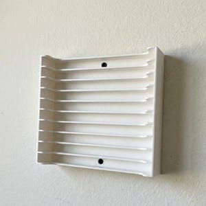 May include: A white, wall-mounted shelf with a slatted design. The shelf has a rectangular shape with rounded corners and two small, black circular accents. The shelf is mounted on a light-colored wall.