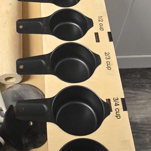 May include: A set of black measuring cups in a wooden organizer. The cups are labeled with standard measurements: 1/8 cup, 1/4 cup, 1/3 cup, 1/2 cup, 2/3 cup, 3/4 cup, and 1 cup. Kitchen tools are visible in the background.