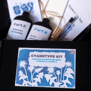 May include: A black box containing a cyanotype kit. The kit includes bottles labeled "Part A" and "Part B", along with paper, a pen, and instructions. The box has a blue and white label that reads "CYANOTYPE KIT experience photography without a camera."