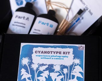 Op de afbeelding: Een zwarte doos met een cyanotypiekit. De kit bevat flessen met het opschrift "Deel A" en "Deel B", samen met papier, een pen en instructies. De doos heeft een blauw en wit label met de tekst "CYANOTYPE KIT experience photography without a camera."