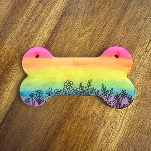 May include: Rainbow-coloured dog bone with a black outline of flowers and leaves.