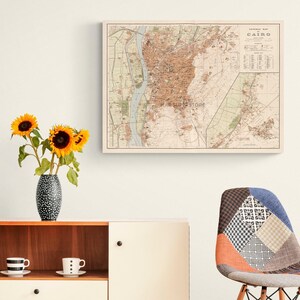 May include: A framed vintage map of Cairo, Egypt, displayed on a wall above a wooden cabinet. The cabinet holds a black and white polka dot vase with three yellow sunflowers and two white coffee cups. A patchwork chair sits to the right.