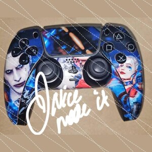 May include: A custom-designed PlayStation 5 controller with a blue and red design featuring the characters Joker and Harley Quinn from the DC Comics universe. The controller has a black background with a blue and red pattern and the characters' faces are prominently displayed on the controller.