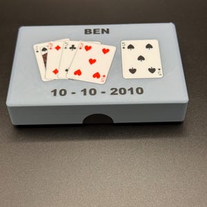 May include: A light blue box with the name "BEN" printed on top. Several playing cards are arranged on the box, including the five of hearts, diamonds, clubs, and spades. The date "10-10-2010" is printed below the cards.