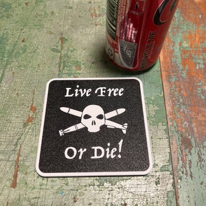Puede incluir: Un posavasos cuadrado negro con texto blanco "Live Free Or Die!" y un diseño de calavera y misiles cruzados. Una lata de refresco roja es visible en el fondo.