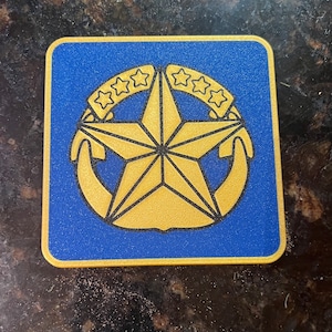 Puede incluir: Posavasos cuadrado azul con un diseño de estrella y ancla dorada. La estrella tiene cinco puntas y está rodeada por un ancla dorada a cada lado. Tres estrellas doradas están encima de la estrella. El posavasos tiene un borde dorado.