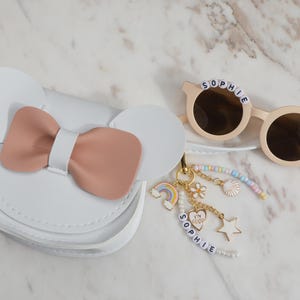 May include: White purse with a pink bow and Mickey Mouse ears. Beige sunglasses with the name "SOPHIE" spelled out in letter beads. A gold keychain with charms, including a rainbow, flower, and star.