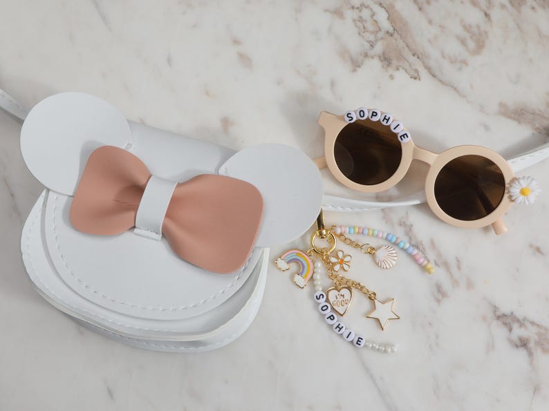 May include: White purse with a pink bow and Mickey Mouse ears. Beige sunglasses with the name "SOPHIE" spelled out in letter beads. A gold keychain with charms, including a rainbow, flower, and star.