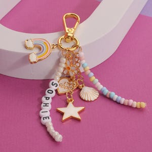 May include: A gold-colored keychain with a rainbow, star, and seashell charms. The keychain has a beaded strand spelling "SOPHIE" and a heart charm that says "I'M GOOD". The background is pink and white.