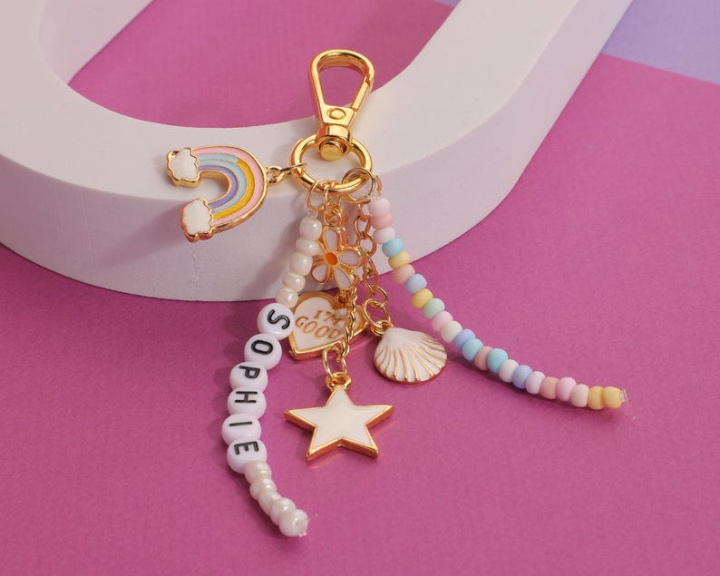May include: A gold-colored keychain with a rainbow, star, and seashell charms. The keychain has a beaded strand spelling "SOPHIE" and a heart charm that says "I'M GOOD". The background is pink and white.