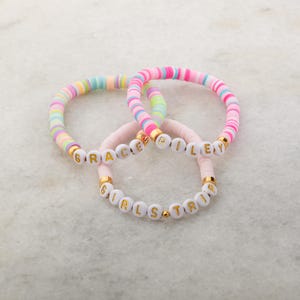 May include: Three colorful beaded bracelets with the names "GRACE", "RILEY", and "GIRLS TRIP" spelled out in gold letters on white beads.
