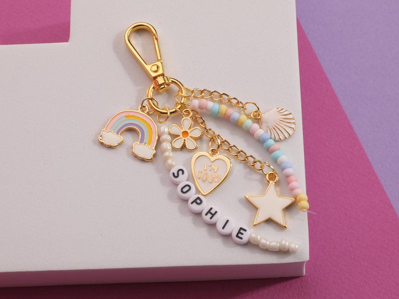 May include: Gold-tone keychain with multiple charms, including a rainbow, flower, heart, star, and seashell. The keychain has a chain and beaded accents. The name "SOPHIE" is spelled out in white letter beads.
